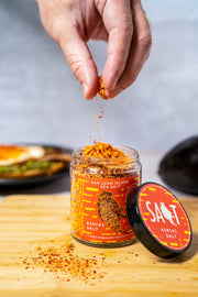 San Juan Island Sea Salt - Kimchi Seasoning Blend spills onto a wooden board, with a hand sprinkling it near avocado toast in the background.