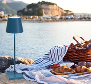 Zafferano America - Poldina Pro Cordless Lamp: Rust enhances a serene waterside picnic, featuring croissants, fruit, and cheese on a striped blanket by the lake.