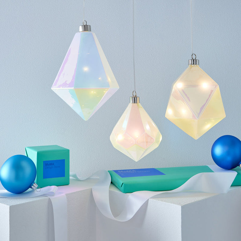 MoMA | LED Glass Prism Ornament: Iridescent, diamond-shaped pendant with warm LED lights, showcased among modern decor elements, adding a festive, ethereal touch to any setting.