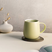 OHOM - Ui 3 Self-heating Mug Set: Jasmine White on electric coaster, beside vase with seed pods, open book edge visible, minimalist aesthetic.