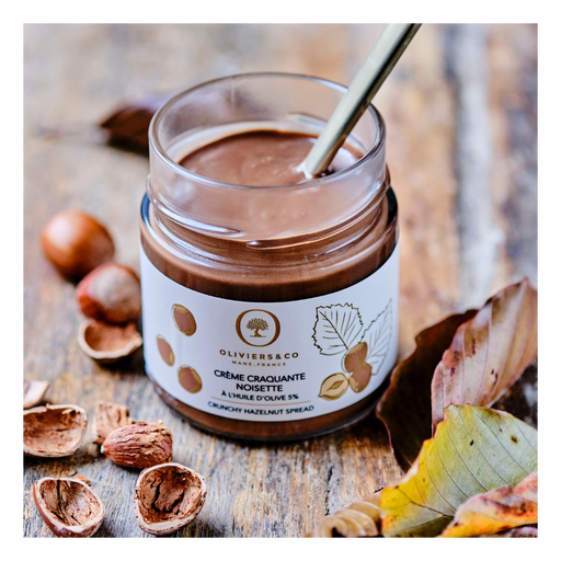 Oliviers&Co | Crunchy Hazelnut Spread in a jar with spoon, surrounded by hazelnuts and shells on a wooden surface.