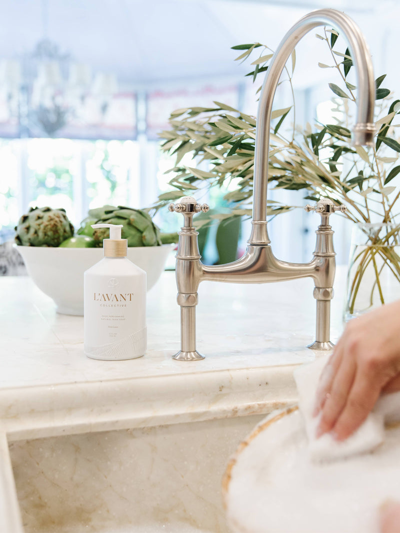L'AVANT Collective Fresh Linen Dish Soap in a matte white glass bottle on a kitchen counter beside a silver faucet and plant.