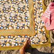 Top-down view of Couleur Nature's Forest Harvest tablecloth, featuring a mustard and blue floral pattern, with a coral towel and a foot visible on the beach.