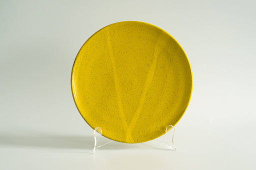 RachaelPots - Dinner Plate: Yellow, a mustard-colored ceramic plate with speckled glaze and subtle V-shape contour, displayed upright on an acrylic stand.