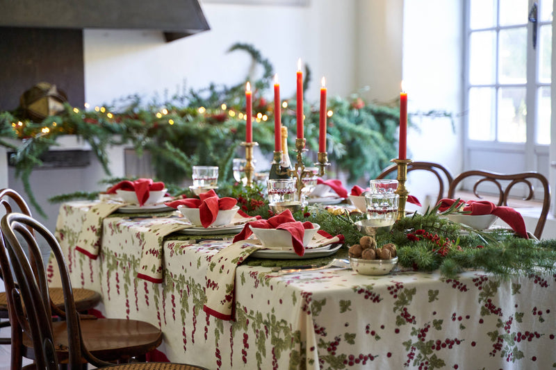 Couleur Nature Gooseberry tablecloth, 71x71, with holly pattern, set with brass candleholders, red tapers, and evergreen garlands for a festive dining table.