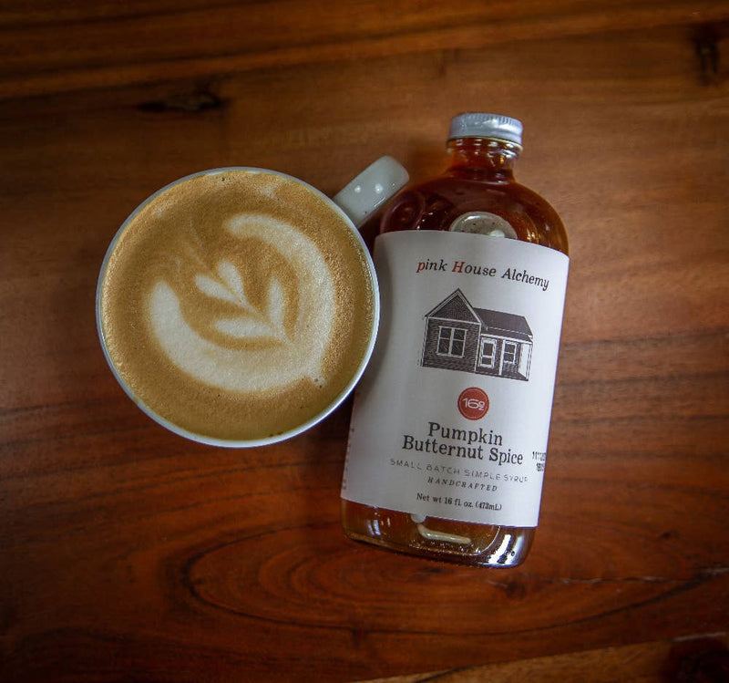 White ceramic mug with latte art beside a 16-oz amber glass bottle of Pink House Alchemy - Pumpkin Butternut Spice Syrup, highlighting warm autumn flavors.