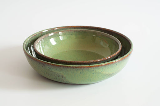 Two green-glazed RachaelPots Pasta Bowls, nestled together, showcasing a speckled finish and brown rims, ideal for serving pasta or noodles.
