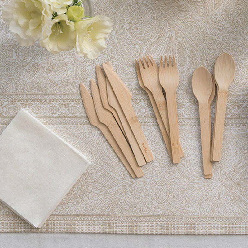 Bambu® Disposable Bamboo Knife, Fork, and Spoon Set displayed on a table with a cream tablecloth, clear vase with flowers, and white napkins, highlighting eco-friendly, compostable design.