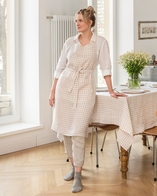 Woman in a natural gingham MagicLinen bib apron, leaning on a table with matching tablecloth, set for dining, in a sunlit dining area.