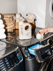 L'AVANT Collective - Laundry Detergent Fresh Linen with measuring cup on modern washer, surrounded by dryer balls, clothespins, and white towels, embodying eco-friendly stain-fighting luxury.