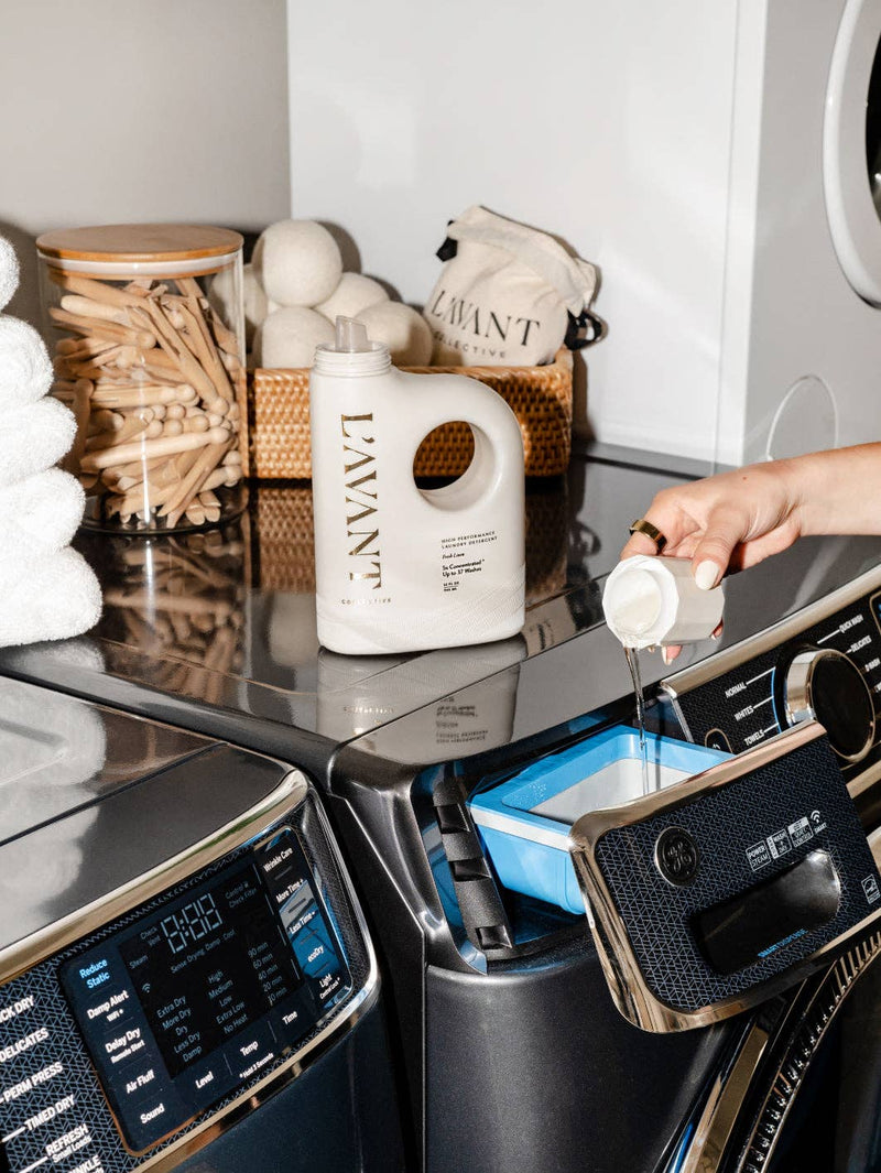L'AVANT Collective - Laundry Detergent Fresh Linen with measuring cup on modern washer, surrounded by dryer balls, clothespins, and white towels, embodying eco-friendly stain-fighting luxury.