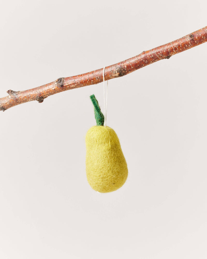 Farmhouse Pottery - Felted Fruit Ornament: Pear, handcrafted in Nepal from yellow wool, hangs by a white string from a tree branch, showcasing its green stem.