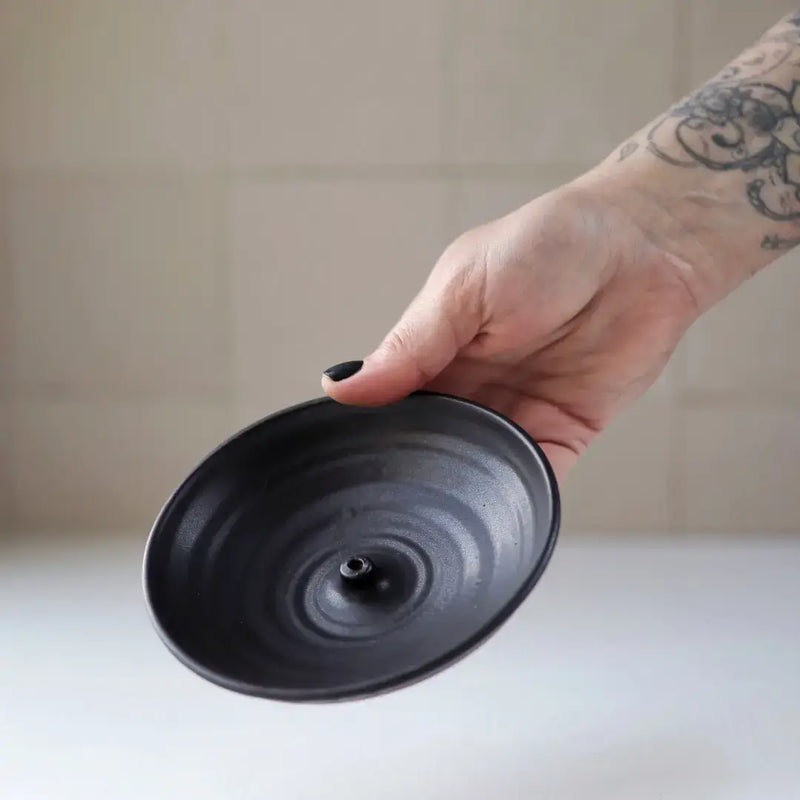 Hand holding Gravesco Pottery - Incense Burner, a black ceramic dish with center peg for incense, ideal for catching ash. Perfect for enhancing kitchen ambiance.