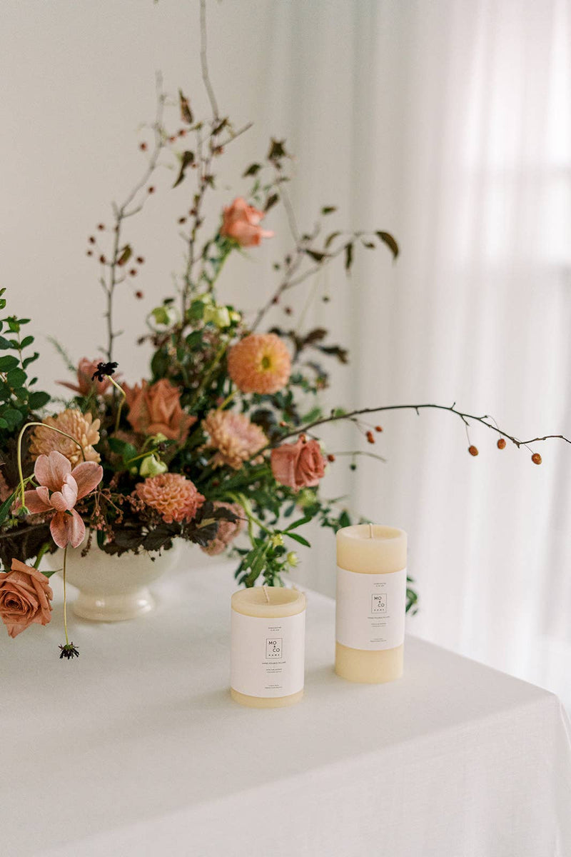 Mo&Co Home - 100% Beeswax Pillar Candles | Natural White: 3x4 Inch, shown beside a vase of flowers, ideal for home decor and celebrations.