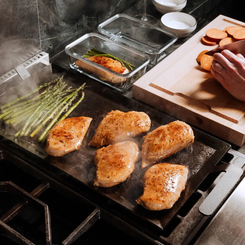 Made In | Carbon Steel Griddle searing chicken and asparagus; sweet potatoes being sliced nearby, showcasing its versatility and heat retention for home cooking.