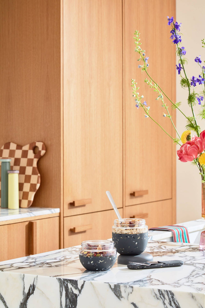 W&P Glass Seal Tight To-Go & Storage Bowl on a marble table, alongside a vase of flowers and a jar of food, showcasing its kitchen utility.