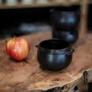 Gravesco Pottery - Black Cauldron Bowl: Handcrafted cast-iron bowl on a wooden slab, ideal for hearty dishes. Accompanied by an apple, showcasing artisanal craftsmanship.