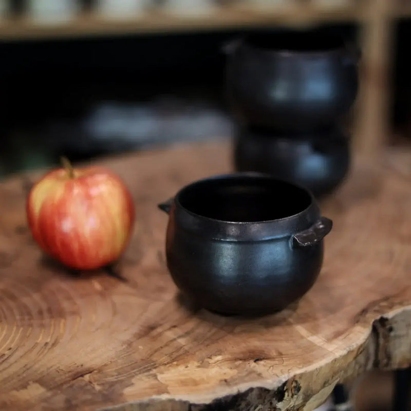 Gravesco Pottery - Black Cauldron Bowl: Handcrafted cast-iron bowl on a wooden slab, ideal for hearty dishes. Accompanied by an apple, showcasing artisanal craftsmanship.