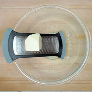 Microplane | Bowl Graters, Fine positioned over a glass bowl with a block of butter, ideal for grating hard cheeses and spices efficiently.