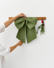 MagicLinen | Linen Decorative Bow, Forest Green, is expertly adjusted on a wooden hook, accompanied by a festive sprig, adding a natural, elegant touch to home décor.