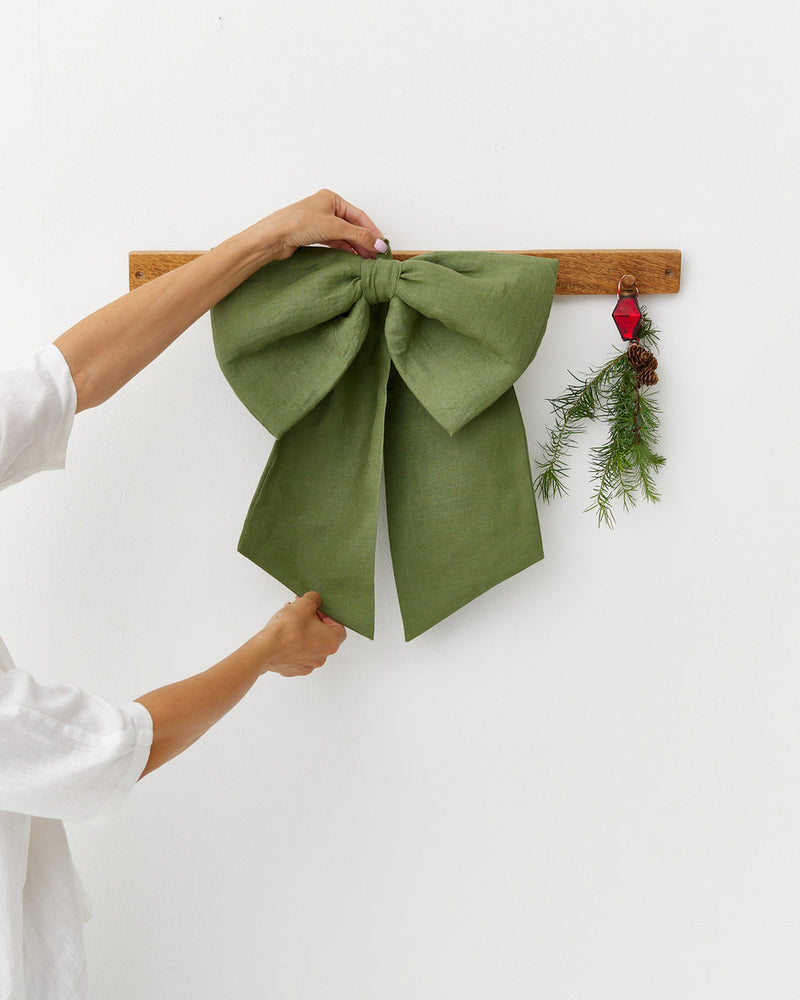 MagicLinen | Linen Decorative Bow, Forest Green, is expertly adjusted on a wooden hook, accompanied by a festive sprig, adding a natural, elegant touch to home décor.