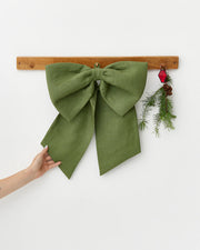 Weston Farms Magnolia Wreath, a lush green and brown wreath, hangs beside an olive-green fabric bow and holiday decor, showcasing festive elegance.