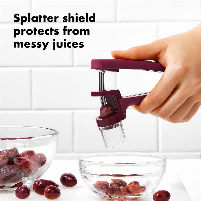 OXO Good Grips Cherry & Olive Pitter in Beet, held by hand, demonstrating pitting functionality over a bowl of olives.