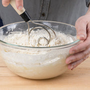 A person using the Brod & Taylor Dough Whisk to mix flour in a glass bowl, ideal for efficiently preparing dough and baking projects.