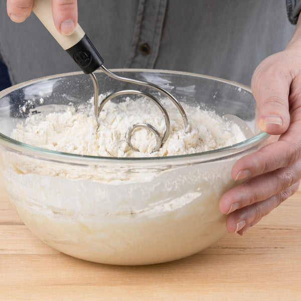 A person using the Brod & Taylor Dough Whisk to mix flour in a glass bowl, ideal for efficiently preparing dough and baking projects.