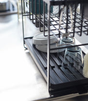 Two-Tier Dish Rack from Yamazaki Home features a black metal frame with a drip tray, drying glasses, plates, and cups on a stainless-steel countertop.