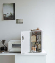 Yamazaki Home vertical steel bread box, black, keeps bread fresh on a minimalist kitchenette with coffee dripper, toaster-oven, and open mini-fridge displaying various foods.