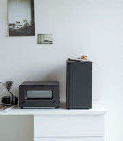Yamazaki Home | Bread Box, Vertical, Steel, Black, sits amid a sleek kitchen setup, enhancing the decor and organizing space, perfect for keeping baked goods fresh.