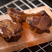 Two seasoned, grilled meat chops rest on the Pink Himalayan Salt Plank with Holder Tray, showcasing its unique cooking and serving capabilities.