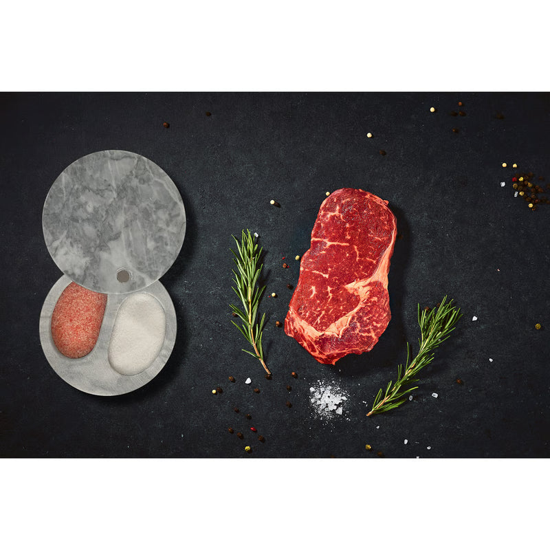 White Marble Double Salt Cellar with pink Himalayan and white sea salt, surrounded by a marbled steak, peppercorns, salt, and rosemary on a dark surface.