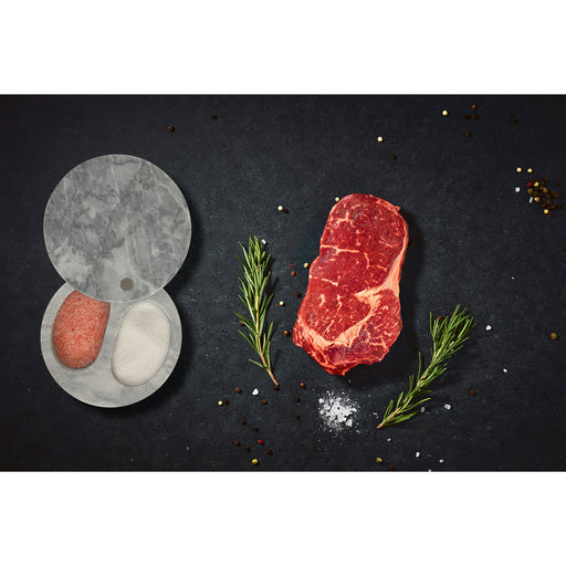 White Marble Double Salt Cellar with pink Himalayan and white sea salt, surrounded by a marbled steak, peppercorns, salt, and rosemary on a dark surface.