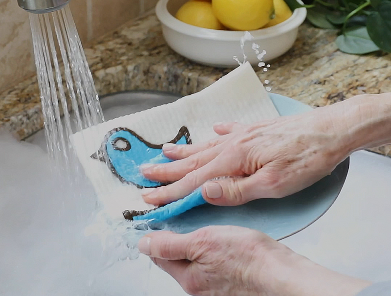 Three Bluebirds Swedish Dishcloths in action; a person washes a plate with a blue towel under running water.