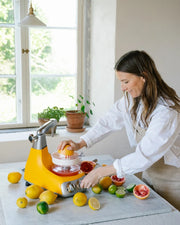 Person using a yellow electric citrus juicer on a sunlit kitchen counter, surrounded by citrus fruits. Product focus: Ankarsrum | Stand Mixers.