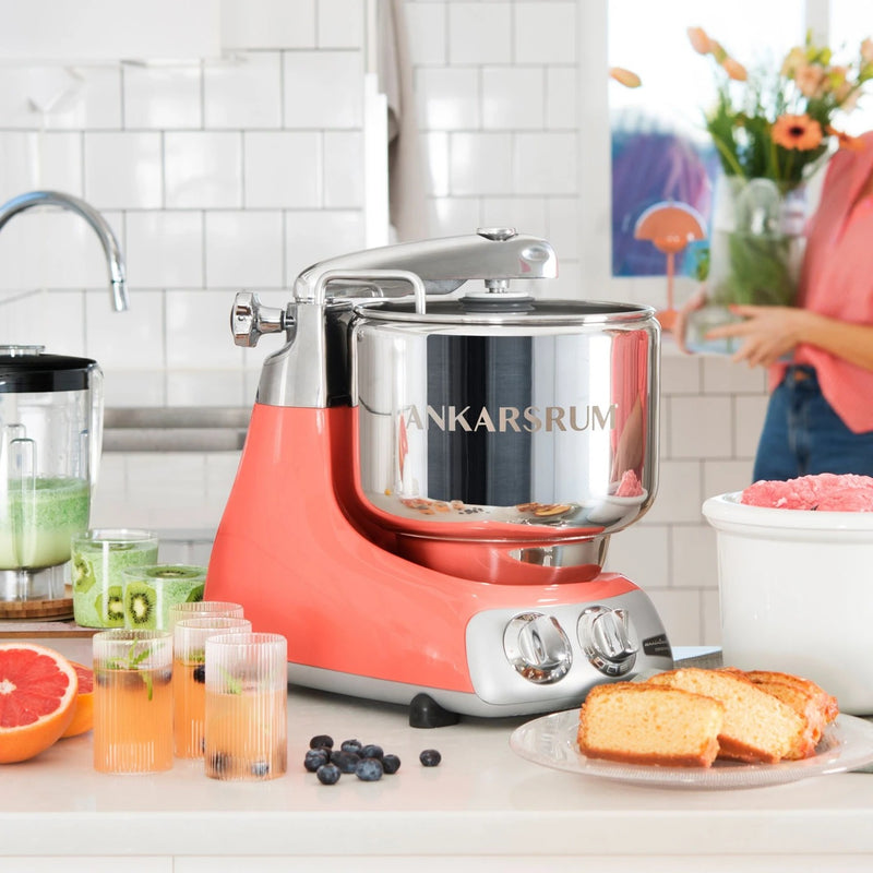 Ankarsrum stand mixer with stainless-steel bowl on countertop, surrounded by a blender, drinks, pound cake, and ice cream; perfect for versatile kitchen creations.