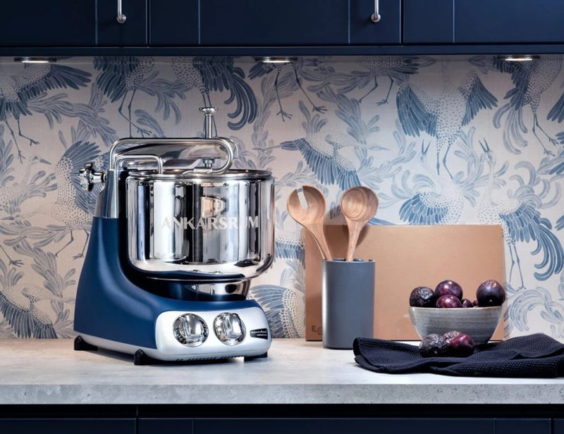 Ankarsrum stand mixer on countertop with utensil crock and plums, against bird-patterned wallpaper.