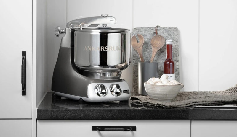 Ankarsrum stand mixer on countertop with control knobs, alongside a cutting board, utensil holder, oil bottle, and a bowl of mushrooms. Ideal for home baking.