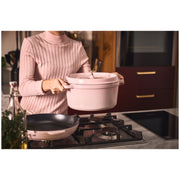 Person holding Staub Round Dutch Oven, Pink Peony, 5.5 QT, above gas cooktop in kitchen, showcasing its elegant design alongside matching skillet and cooking essentials.