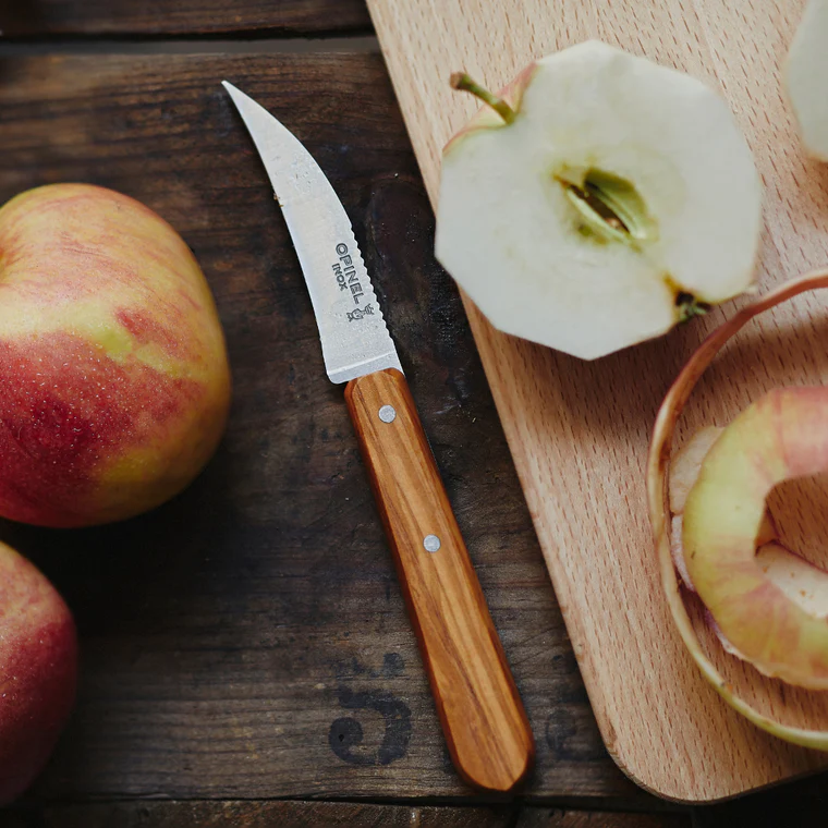Opinel Essential Birdsbeak Vegetable Knife on a dark wooden surface with apple peels and cores, showcasing its precise cutting ability for fruits and vegetables.
