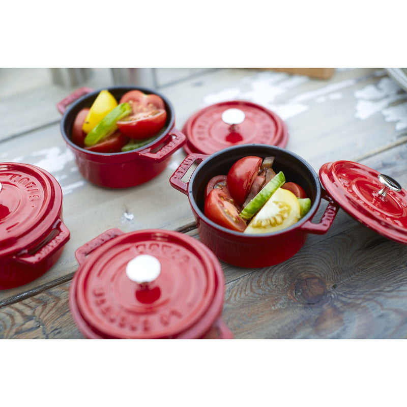 Staub Mini Round Dutch Oven, Cherry, 0.275 QT, displayed on a rustic table, with open pots revealing vibrant heirloom tomatoes and cucumbers, showcasing culinary elegance.