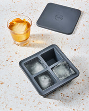 W&P | Peak Reusable Silicone Ice Trays - Charcoal Extra Large, showing three large frozen cubes, one empty spot, and a whisky glass with a cube and orange peel.