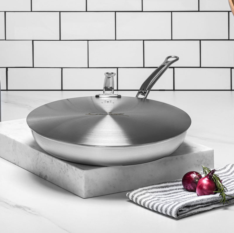 Hestan Commercial Clad Stainless Steel Universal Lids, 12.5 Inches, on marble with towel, red onions, and rosemary; steam vent reduces pressure and prevents boil overs.