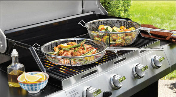 A Fox Run Brands 3 in 1 Grilling Basket filled with a variety of grilled vegetables and meat sits outdoors. The grill, ideal for both indoor and outdoor cooking, features multiple knobs and a side tray holding a blue and white bowl of lemon wedges. A bottle of oil is also on the tray, with fresh green bushes in the background.