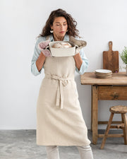 Woman holding a baking dish with a loaf of bread, featuring a MagicLinen Linen pot-holder in Woodrose, showcasing heat-insulating padding for kitchen use.