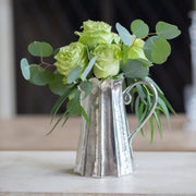 Arte Italica & Crown Linen Designs Vintage Tall Scalloped Pitcher, crafted in Italian pewter, serves as a vase for roses and eucalyptus on a tabletop.