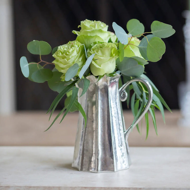 Arte Italica & Crown Linen Designs Vintage Tall Scalloped Pitcher, crafted in Italian pewter, serves as a vase for roses and eucalyptus on a tabletop.