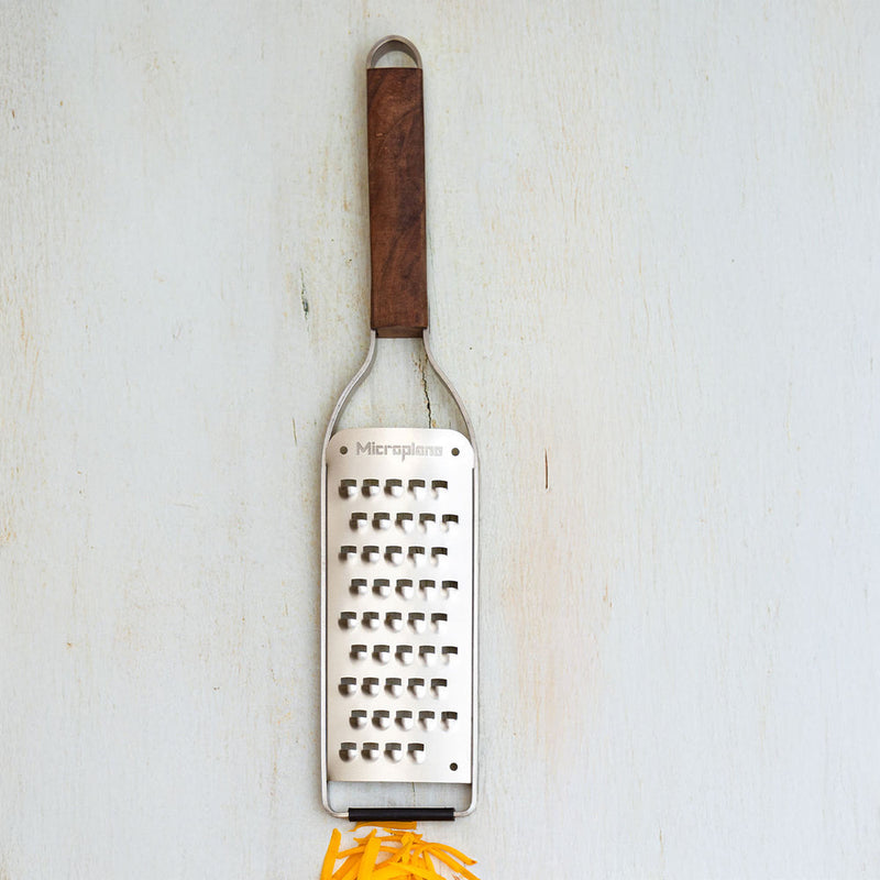 Microplane | Master Series Extra Coarse Grater with Walnut Handle, featuring a stainless-steel blade and wooden handle, shown with freshly shredded cheese beneath it.